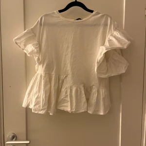 Vit volangblus från Zara - Säljer en vit blus från Zara i storlek M. Blusen har korta, voluminösa ärmar och bred volang nedtill som ger en cool och luftig look. Rund halsringning och tillverkad i mjuk bomull. Perfekt för dig som gillar romantiska detaljer och oversized känsla.