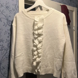 Vit stickad tröja med volangdetalj - Supersöt vit stickad tröja med lång ärm och bred volang framtill som ger en extra touch. Rund halsringning och ribbade muddar vid ärmslut och nederkant. Perfekt för dig som gillar en clean look med lite extra detalj.
