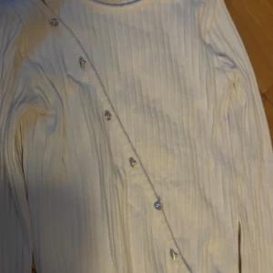 Vit ribbad kofta med glittriga knappar - Säljer en vit ribbad kofta med lång ärm och snygga glittriga knappar i silver längs framsidan. Koftan har en cool asymmetrisk knäppning och är tillverkad i mjukt material som känns skönt mot huden. Perfekt för dig som gillar stilrena detaljer.