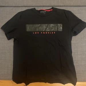 Svart GUESS t-shirt med tryck - Svart t-shirt från GUESS med stilrent tryck på bröstet där det står 'GUESS' i svart och 'LOS ANGELES' i rött. Klassisk passform och rund halsringning. Tillverkad i mjuk bomull, perfekt för en clean och modern look.