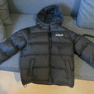 Svart pufferjacka från Hoodrich - Svart pufferjacka från Hoodrich med stor logga på ryggen och mindre logga på bröstet. Jackan har huva, dragkedja framtill och två sidofickor. Materialet är syntet och jackan har en quiltad design som ger en fet och puffig look. Perfekt för kalla dagar.