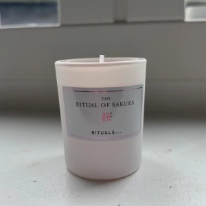 The Ritual of Sakura Doftljus - Elegant doftljus från Rituals, The Ritual of Sakura. Ljusrosa vax i vit behållare, oanvänt med hel veke. Ger en fräsch och avslappnande doftupplevelse. Perfekt för hemmet eller som present. Inga synliga defekter eller slitage. Helt oanvänd.