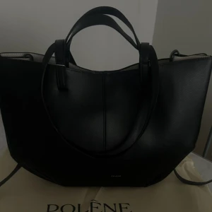 Svart handväska från Polène - Stilren svart handväska från Polène med elegant och modern form. Väskan har både korta och långa handtag, vilket gör den enkel att bära på olika sätt. Tillverkad i slätt skinn med minimalistisk design och diskret logga framtill.