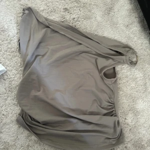 Beige off shoulder topp med ringdetalj - Snygg beige topp med en cool ringdetalj i guld på ena axeln. Toppen har en enkel och stilren design, stretchigt material som sitter skönt. Perfekt för dig som gillar minimalistisk stil och vill sticka ut lite.