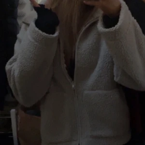 Beige teddyjacka med dragkedja - Mysig beige teddyjacka med dragkedja framtill och stora fickor. Jackan har en avslappnad passform och är perfekt för kyliga dagar. Den fluffiga ytan ger en trendig look och är verkligen superfin