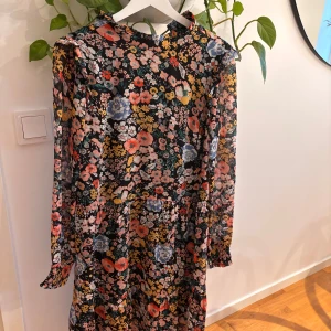 Blommig långklänning från Co'couture - Snygg långklänning från Co'couture med färgglatt blommigt mönster i rosa, blått, gult och grönt på svart botten. Klänningen har långa ärmar med lätt rynk vid mudden och en rund halsringning. Perfekt för dig som gillar statement-plagg med mycket färg.