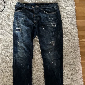 Mörkblå jeans från Alessandro Zavetti - Snygga mörkblå jeans från Alessandro Zavetti med slitningar, hål och färgstänk för en riktigt edgy look. Klassisk femficksmodell med raka ben och coola detaljer. Perfekta för dig som gillar streetstyle och vill sticka ut lite extra.