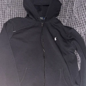 Svart hoodie från Polo Ralph Lauren - Svart hoodie från Polo Ralph Lauren med dragkedja och huva. Klassisk design med vit broderad logga på bröstet, snörning vid huvan och känguruficka framtill. Tillverkad i mjuk bomullsmix som är skön att ha på sig. Perfekt för chill dagar.