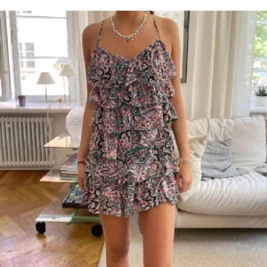 Blommig kortklänning med volanger - Supersöt kortklänning med blommigt mönster i rosa, svart och grönt. Klänningen har tunna axelband som korsas i ryggen, volanger och en öppen rygg. Perfekt för sommarens alla tillfällen och ger en riktigt fräsch vibe.