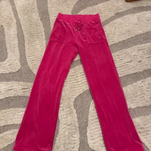 Juicy Couture rosa mjukisbyxor S - Rosa mjukisbyxor från Juicy Couture med bred resår i midjan och snörning framtill. Byxorna har stora fickor både fram och bak, samt broderad JC-logga på ena fickan. Mjukt och glansigt material som känns lyxigt och skönt.