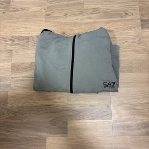 Grå hoodiejacka EA7 Emporio Armani - Snygg grå hoodiejacka från EA7 Emporio Armani med svarta detaljer längs ärmarna och dragkedja framtill. Jackan har huva med snörning och två fickor fram. Perfekt för chill eller sportiga dagar.