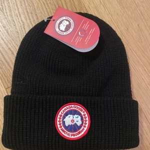 Svart Canada Goose mössa - Svart ribbstickad mössa från Canada Goose med klassisk rund patch framtill i rött, vitt och blått. Mössan har uppvikt kant och är tillverkad i ett tjockt, stickat material som håller dig varm även när det är riktigt kallt ute. Perfekt för kalla vinterdagar.