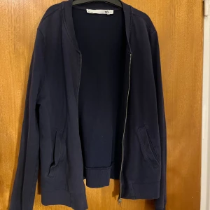 Mörkblå bomberjacka från 157 - Snygg mörkblå bomberjacka från 157 med klassisk ribbad krage och muddar. Jackan har två fickor framtill och stängs med dragkedja. Tillverkad i mjuk bomull som är skön att ha på sig. Perfekt för dig som gillar enkel och stilren design.