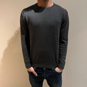 Mörkgrå stickad tröja  - En stilren mörkgrå långärmad tröja med rund hals. Tröjan har en enkel design utan mönster och är perfekt för dig som gillar en clean look. Materialet känns mjukt och skönt mot huden, och passformen är normal med ribbade muddar vid ärmslut och nederkant.