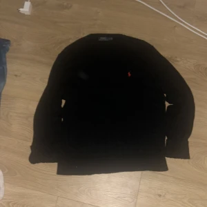 Svart stickad tröja från Polo Ralph Lauren - Svart stickad tröja från Polo Ralph Lauren med klassisk kabelstickad design och röd broderad logga på bröstet. Tröjan har rund hals och långa ärmar, perfekt för dig som gillar stilrena och tidlösa plagg.