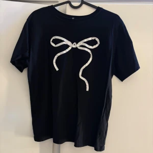 Svart t-shirt med rosettdetalj H&M - Svart t-shirt från H&M med en vit rosett framtill. Sitter snyggt och skönt på kroppen!