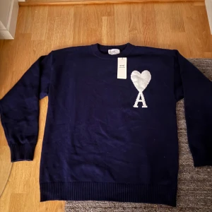 Marinblå stickad tröja från Ami Paris - Passar stor XS. Snygg marinblå stickad tröja från Ami Paris med stort vitt hjärta och bokstaven A på bröstet. Tröjan har rund hals, ribbade muddar och lång ärm. Perfekt för dig som gillar stilrena detaljer och vill ha något som sticker ut lite extra.