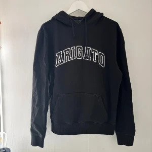 Svart hoodie från Axel Arigato - Svart hoodie från Axel Arigato med vit broderad logga på bröstet. Klassisk huva med snörning och stor magficka framtill. Långärmad och i mjuk bomull, perfekt för chill dagar. Snygg och enkel stil som funkar året runt.