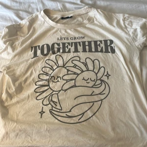 Vit t-shirt med tryck från LMTD - Säljer en vit t-shirt från LMTD i storlek M med ett gulligt tryck på framsidan som visar två tecknade blommor som kramas och texten 'Lets Grow Together'. T-shirten har rund hals och korta ärmar, perfekt för dig som gillar söta och positiva motiv. 