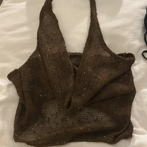 Brun glittrig croppad linnetopp - Säljer en brun croppad linnetopp med glittriga paljettdetaljer. Toppen har djup omlott-ringning och är ärmlös med breda axelband. Materialet är stickat och ger en cool mesh-effekt. Perfekt för att sticka ut med lite extra shine. (Ganska genomskinlig topp med djup rygg! Så fin till middag eller temafest! Aldrig använd!)