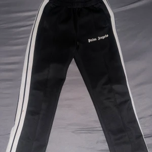 Palm Angels svarta trackpants med vita ränder - Svarta trackpants från Palm Angels med vita sidoränder och logga på vänster ben. Byxorna har elastisk midja med snörning och dragkedjor vid bensluten. Tillverkade i glansig polyester för en sportig vibe.