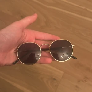 Runda solglasögon med guldram - Snygga runda solglasögon med tunn guldfärgad metallbåge och mörka glas. Skalmarna är svarta och har en klassisk design. Perfekta för att lyfta din stil under soliga dagar.