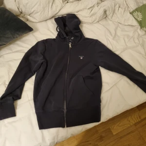 Svart hoodie med dragkedja från GANT - Svart hoodie från GANT med broderad logga på bröstet. Tröjan har huva med snörning, hel dragkedja framtill och två fickor. Tillverkad i mjuk bomullsblandning, perfekt för chill dagar. Klassisk och stilren design som funkar året runt.