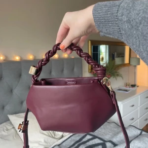 Vinröd handväska med flätat handtag - Frågan är som den ör på första fyra bilderna,mörkt ljus i sista bilden. Snygg vinröd handväska i skinn med unik flätad detalj på handtaget och guldfärgade metalldetaljer. Väskan har en avtagbar axelrem och en stilren, geometrisk form som gör den till ett statement piece. Perfekt storlek för det viktigaste och stängs med magnetknapp.