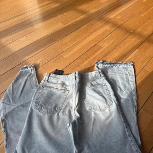 Ljusblå bootcut jeans med rå kant - Säljer ett par ljusblå jeans med flared passform och rå, fransig kant längst ner. Jeansen har klassisk femficksdesign och är tillverkade i jeansmaterial. Färgskiftningar och små vita stänk ger en cool, vintage vibe.