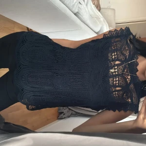 Svart spetsblus med detaljerad design - Mörk blå blus med spetsmönster och genombrutna detaljer vid halsen. Blusen är ärmlös och har en feminin, elegant look med vackra kanter och hög hals.