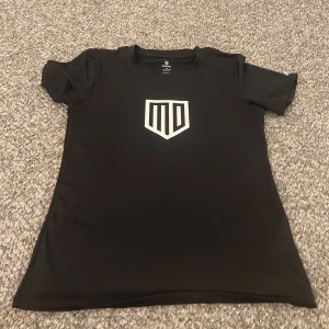 Svart t-shirt från Maya Delorez - Svart t-shirt från Maya Delorez med vit logga framtill. Klassisk rund hals och korta ärmar. Tillverkad i ett mjukt och stretchigt syntetmaterial som andas, perfekt för en aktiv livsstil.