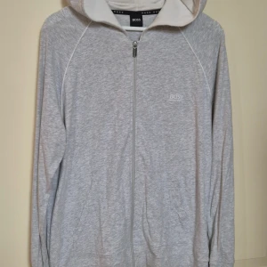 Grå hoodie från Hugo Boss med dragkedja L - Snygg ljusgrå hoodie från Hugo Boss i storlek L. Tröjan har hel dragkedja framtill, huva med vit insida och diskret Boss-logga på bröstet. Tillverkad i mjuk bomullsmix, perfekt för chill eller när du vill ha en clean look. Fickor på sidorna och långa ärmar. I fint skick!