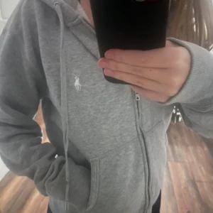 Grå hoodie från Polo Ralph Lauren - Säljer en grå hoodie från Polo Ralph Lauren i storlek M. Tröjan har dragkedja, snörning vid huvan och två fickor framtill. Klassisk vit logga broderad på framsidan. Använd men inga skador på tröjan! Säljer för att den är för stor och aldrig kommer till användning🫶