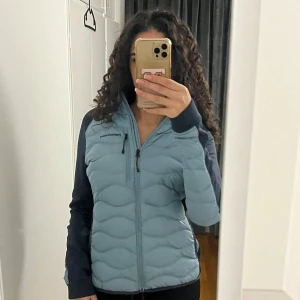 Blå pufferjacka Peak Performance - Snygg blå pufferjacka från Peak Performance med svarta ärmar och huva. Jackan har quiltad framsida, dragkedja hela vägen och en bröstficka med dragkedja. Perfekt för kyliga dagar och har en sportig vibe. Storleken är XL, men skulle säga att det passar nån med stil M/L. För referens har jag S i jackor vanligtvis.