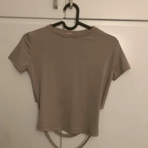 Beige croppad topp med öppen rygg - Säljer en beige croppad topp med korta ärmar och rund hals. Toppen har snygga snörningsdetaljer i nederkant (rygg) som ger en trendig look. Materialet känns mjukt och stretchigt, perfekt för dig som gillar stilrena och enkla plagg med en twist.