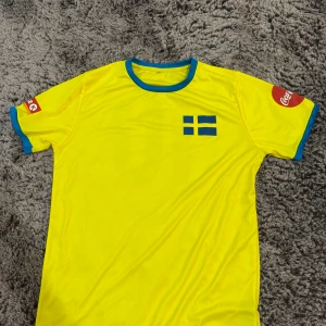 Svensk fotbollströja XL med nummer 10 - Gul fotbollströja i polyester med blå detaljer och svensk flagga på bröstet. Nummer 10 på ryggen och Coca-Cola-logga på båda ärmarna. Rund hals med blå kant och blå muddar vid ärmslut. Perfekt för dig som vill sticka ut på matchen.
