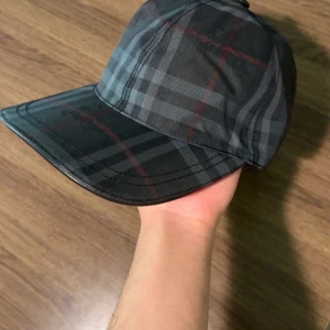 Burberry rutig keps i mörka toner - Snygg Burberry keps med klassiskt rutigt mönster i mörkgrått, svart och röda linjer. Justerbar rem baktill med metallspänne. Materialet är bomull och polyester, perfekt för dig som vill ha en stilren och lyxig look. Passar dig som gillar designeraccessoarer.