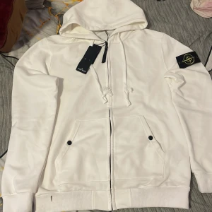 Vit hoodie från Stone Island med huva - Vit hoodie från Stone Island med klassisk logotyp på ärmen, dragkedja framtill och två fickor med knappar. Tröjan har huva med snörning och är tillverkad i mjuk bomull. Perfekt för dig som gillar clean och stilren streetwear.