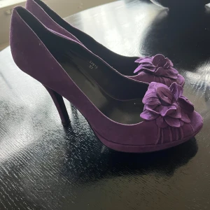 Snygga skor  från Office - Säljer ett par lila pumps från Office i mocka med spetsig tå och hög klack. Skorna har en snygg blomdetalj framtill som ger en extra touch. Perfekta för dig som vill sticka ut med färg och stil. Köpt i London