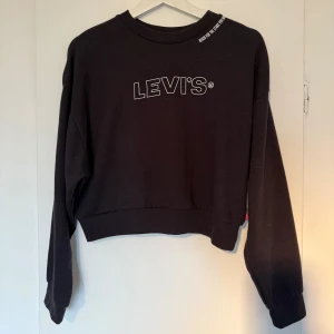 Svart croppad sweatshirt från Levi's - Svart croppad sweatshirt från Levi's med vit logga framtill och tryck på ena ärmen. Tröjan har rund halsringning och långa ärmar