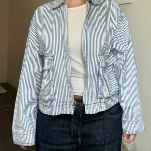 Randig jeansjacka från H&M Divided - Säljer en snygg randig jeansjacka från H&M Divided i storlek S. Jackan är vit med blå vertikala ränder, har dragkedja framtill, två fickor med knapp och klassisk krage. Materialet är tyg och passformen är normal med långa ärmar. Jackan är använd några enstaka gånger bara <3