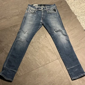 Replay Anbass jeans blå, W32 L30 - Säljer ett par Replay Anbass jeans i klassisk blå tvätt med snygga slitningar. Modellen har fem fickor, raka ben och en smal passform. Jeansen är tillverkade i bomull med en skön stretch och har detaljerade sömmar på bakfickorna.