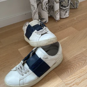 Valentino Garavani Open sneakers vit/blå - Snygga Valentino Garavani Open sneakers i vitt skinn med bred blå läderdetalj över vristen. Klassisk rund tå, platt sula och grå snörning. Diskret Valentino-logga på plösen och nitar baktill. Lite tecken på användning under skon då den är relativt sliten under men annars inga problem på skicket. Perfekta för dig som gillar lyxiga och stilrena sneakers. Pris går att diskutera!!