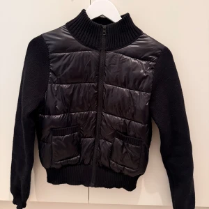 Svart pufferjacka från Gina Tricot - Svart pufferjacka med stickade ärmar och ribbad krage från Gina Tricot. Framsidan har quiltade paneler i blankt syntetmaterial och två fickor med ribbade detaljer. Jackan är croppad och har dragkedja framtill. Perfekt för dig som gillar en cool och modern look. Använd fåtal gånger, så i jättefint skick! Gina tricot young, men passar mig med storlek M. 