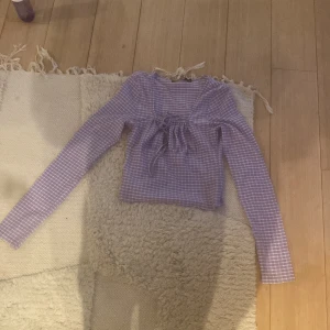 Lila rutig topp med snörning från Bik Bok - Superfin lila långärmad topp från Bik Bok i storlek XS. Toppen har ett rutigt mönster i vitt och lila, croppad passform och snygg snörning framtill vid bysten. Perfekt att styla med jeans eller kjol för en trendig look.