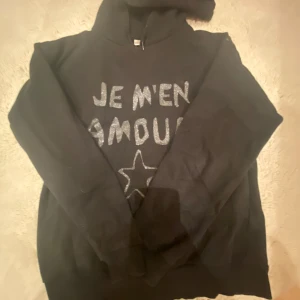 Svart hoodie med glittertext från Saltroot - Svart hoodie från Saltroot med glittrig text 'Je m'en amour' och en stjärna framtill. Tröjan har huva och långa ärmar, perfekt för dig som gillar en chill och snygg look. Materialet är mjuk bomull och passformen är normal.
