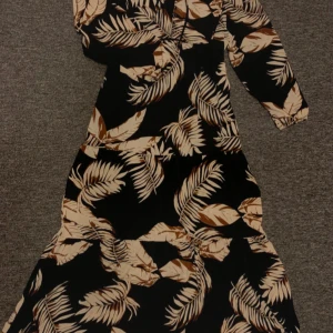 Svart långklänning med beige palmblad - Långklänning med svart botten och stora beige palmbladsmönster. Klänningen har långa ärmar, v-ringning och snörningsdetalj framtill. Luftig och flowy modell som ger en avslappnad men ändå snygg look.
