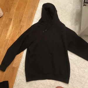 Svart hoodie  - Svart hoodie från DenimDay med stilren design och diskret logga på bröstet. Tröjan har huva med snörning, lång ärm och en stor magficka framtill. Mjuk bomullskvalitet perfekt nu till vintern