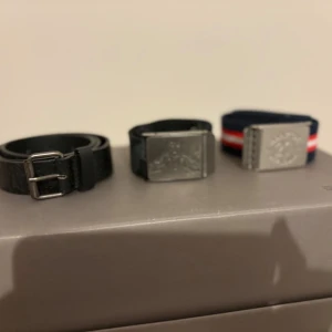 3-pack bälten: svart, randigt & tryckt - Säljer tre bälten: ett svart bälte i skinn med klassiskt spänne, ett svart tygbälte med tryck och metallspänne samt ett marinblått tygbälte med röda och vita ränder och metallspänne. Perfekt för att variera din stil och matcha olika outfits.