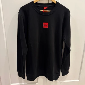 Svart långärmad tröja Hugo Boss - Svart långärmad tröja från Hugo Boss med rund hals och röd Hugo-logga på bröstet. Tillverkad i 100% bomull för skön och mjuk känsla. Stilren design med ribbade muddar vid ärmslut och nederkant. Perfekt för dig som gillar minimalistisk och clean look.